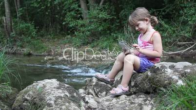 little girl sitting next to a stream and play with tablet