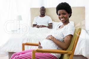 Smiling pregnant woman sitting on chair and man sitting on bed