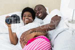 Pregnant couple lying on bed and looking photos in camera