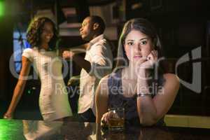 Unhappy woman sitting at bar counter and couple dancing behind h