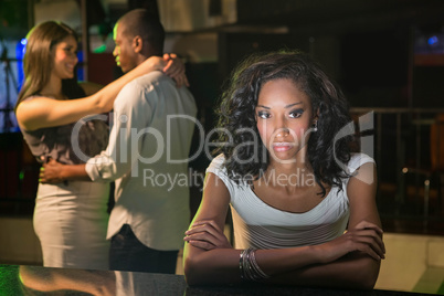 Unhappy woman sitting at bar counter and couple dancing behind h