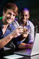 Two men toasting their whiskey glasses at bar counter while usin