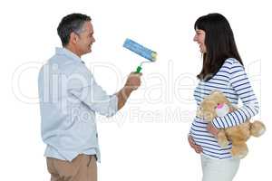 Pregnant woman holding teddy bear while man holding paint roller
