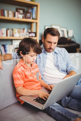 Serious father and son using laptop while sitting on sofa