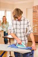 Handsome man ironing a shirt while girlfriend watching in backgr