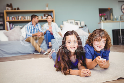 Happy children lying on carpet while parents in background