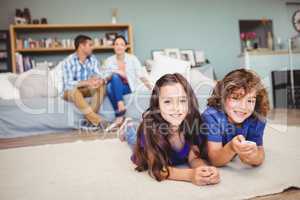Happy children lying on carpet while parents in background