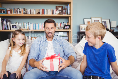 Smiling father holding gift box with children sitting on sofa