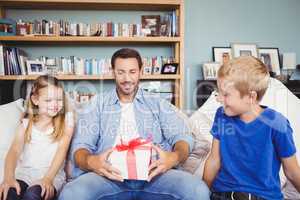 Smiling father holding gift box with children sitting on sofa