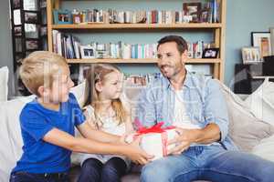 Children giving gift box to father while sitting on sofa
