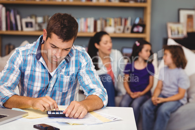 Serious man using calculator while family sitting in background