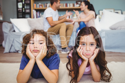 Sad siblings lying on carpet while parents sitting in background