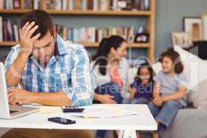 Tensed man by laptop while family sitting in background