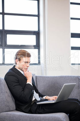 Thoughtful businessman sitting on sofa and using laptop