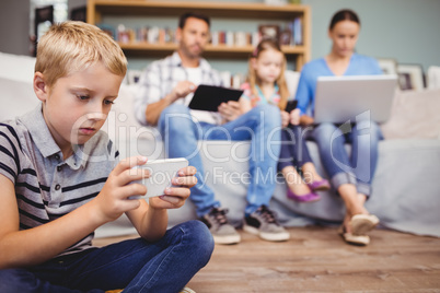 Boy using mobile phone while family with technologies in backgro