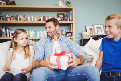 Happy father holding gift box with children sitting on sofa