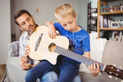 Father teaching son to play guitar while sitting on sofa