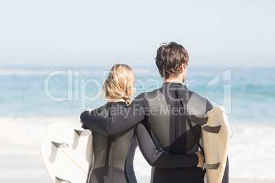 Rear view of couple in wetsuit with surfboard standing on the be