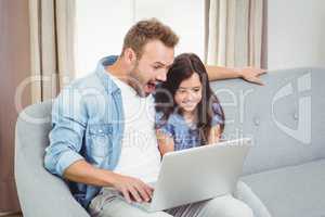 Surprised father and daughter using laptop while sitting on sofa
