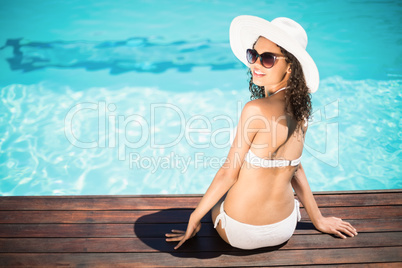 Beautiful woman wearing white bikini and hat sitting near pool