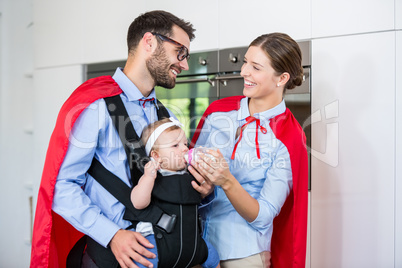 Cheerful couple in superhero costume feeding milk to daughter