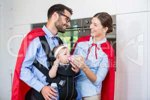 Cheerful couple in superhero costume feeding milk to daughter