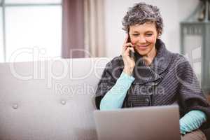 Mature woman with laptop talking on mobile phone at home