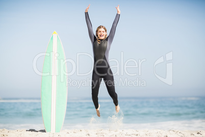 Excited woman in wetsuit jumping next to her surfboard