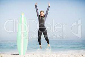 Excited woman in wetsuit jumping next to her surfboard