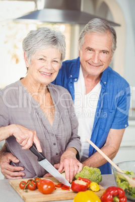 Portrait of senior woman cutting while man embracing in kitchen
