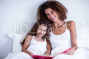 Portrait of mother and daughter reading book together on bed