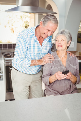 Happy senior man standing besides wife sitting in kitchen