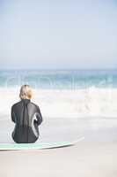 Rear view of woman in wetsuit sitting with surfboard on the beac
