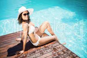 Beautiful woman wearing white bikini and hat sitting near pool