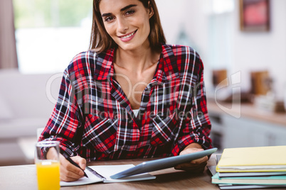 Young woman writing in notepad while holding digital tablet