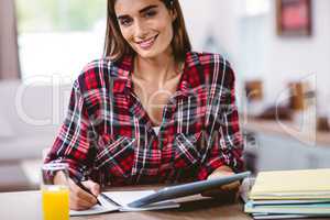 Young woman writing in notepad while holding digital tablet