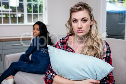 Sad woman holding pillow while female friend sitting on sofa