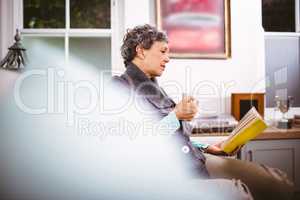 Mature woman reading book and holding coffee mug while sitting o