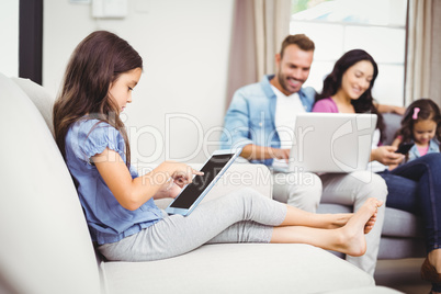 Girl using digital tablet on sofa while family in background