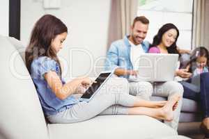 Girl using digital tablet on sofa while family in background