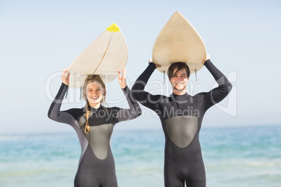 Portrait of couple in wetsuit carrying surfboard over head