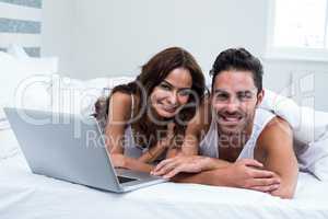 Portrait of smiling couple with laptop while lying under blanket