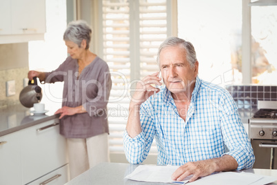 Senior man talking on mobile phone with wife making tea in backg