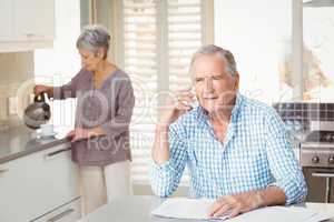 Senior man talking on mobile phone with wife making tea in backg