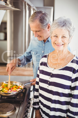 Portrait of senior woman standing with husband cooking food in b