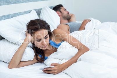 Woman using phone while relaxing on bed husband in background