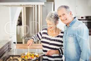 Portrait of senior man standing with wife cooking food
