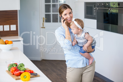 Woman talking on cellphone carrying daughter by kitchen counter