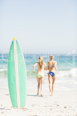 Surfboard in sand and two rear women running on the beach