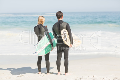 Rear view of couple in wetsuit with surfboard standing on the be